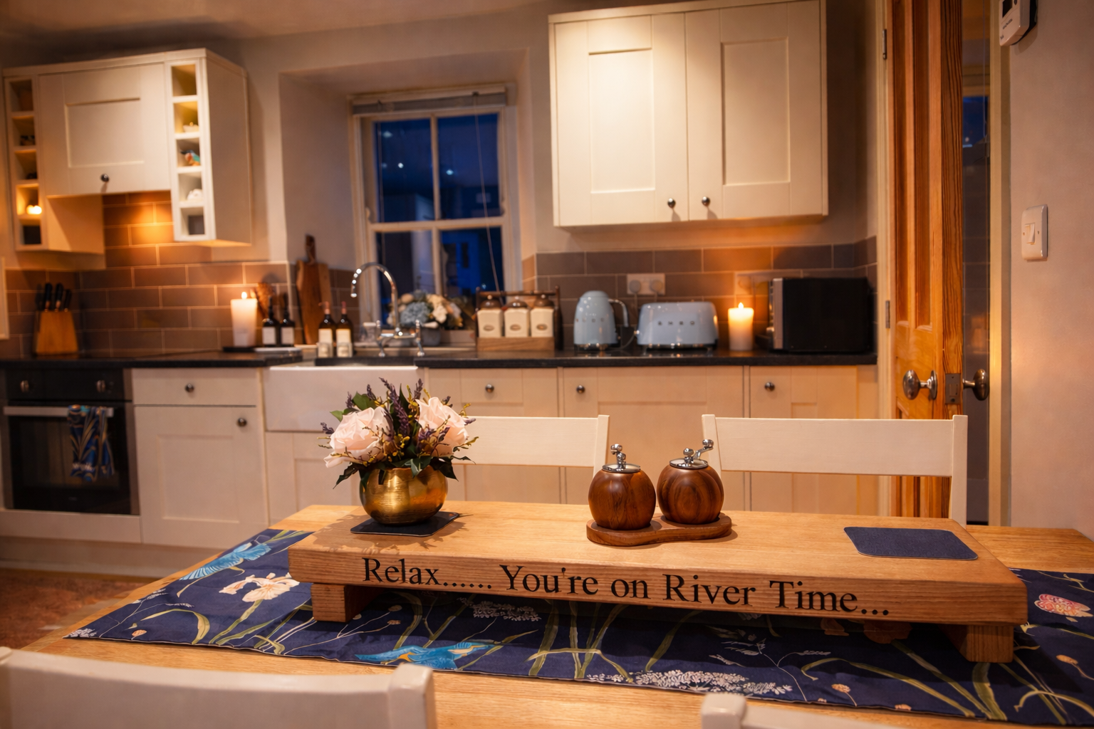 Candlelit cream kitchen and dining table, with grey tiled splashbacks, a built-in oven, sink, toaster, flowers and a wooden River Time board